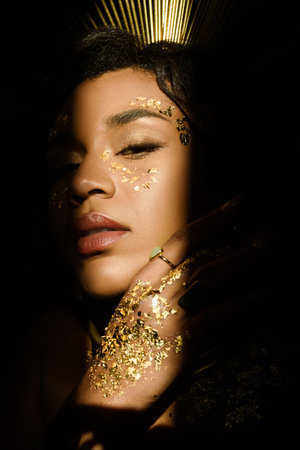 young african american woman with golden accessories and paint on face looking at camera through...