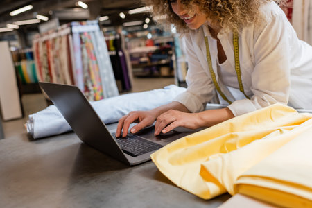 cropped view of happy saleswoman smiling and using laptop near fabric rolls in textile shop