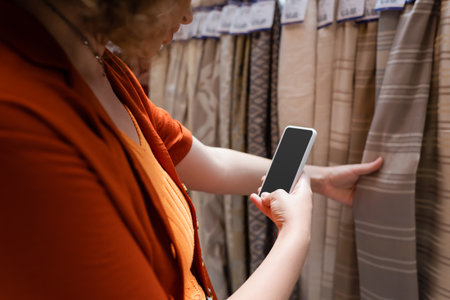 Cropped view of young woman using smartphone with blank screen near fabric in textile shopの写真素材