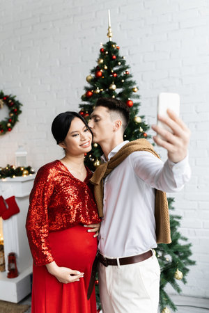 young man kissing elegant and pregnant asian wife while taking selfie near blurred Christmas treeの写真素材