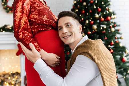 pleased man leaning on tummy of pregnant wife in red shiny blouse during Christmas celebrationの写真素材