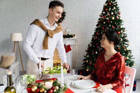 smiling man serving meal near pregnant asian wife sitting at table with romantic Christmas supperの写真素材