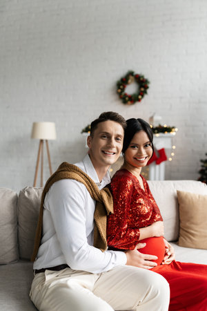 cheerful man with pregnant asian wife in festive outfit looking at camera on couch at homeの写真素材