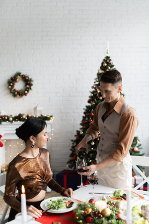elegant man pouring red wine near asian wife in festive clothes during romantic Christmas supper at homeの写真素材