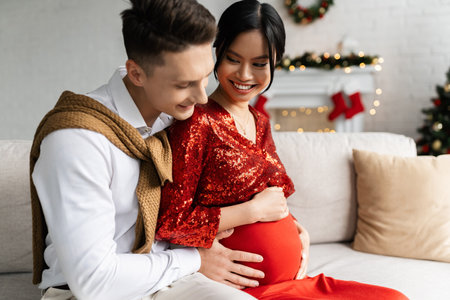 pregnant and elegant asian woman smiling near husband touching her tummy on sofa in living room with Christmas decorの写真素材