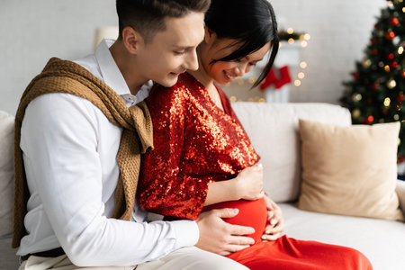 young man hugging tummy of pregnant and happy asian woman while celebrating Christmas at homeの写真素材