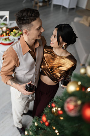 high angle view of happy interracial couple in festive outfits holding glasses of wine near decorated Christmas treeの写真素材