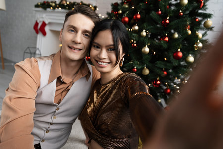 interracial couple smiling and looking at camera while sitting under Christmas treeの写真素材