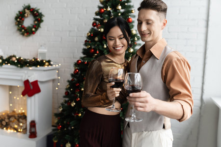 smiling multiethnic couple holding glasses of red wine near Christmas treeの写真素材
