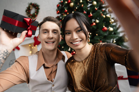 happy tattooed man holding present near asian wife while sitting under Christmas treeの写真素材