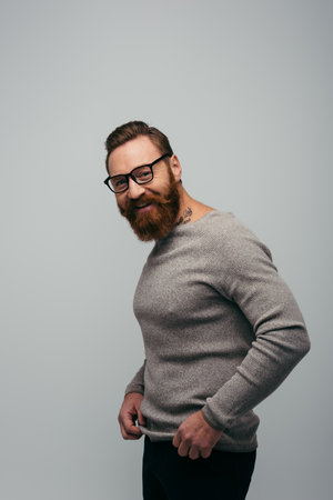 Cheerful bearded man in eyeglasses adjusting jumper and looking at camera isolated on greyの写真素材