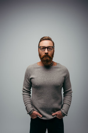 Stylish bearded man in jumper holding hands in pockets of pants isolated on greyの写真素材