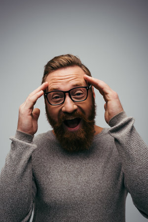 Excited and bearded man in eyeglasses looking at camera isolated on greyの写真素材