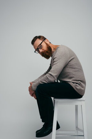 Stylish man in jumper and eyeglasses sitting on chair isolated on greyの写真素材