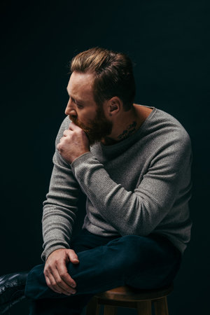 Side view of stylish tattooed man in grey jumper sitting on chair isolated on blackの写真素材