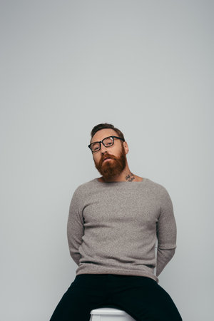Trendy bearded man in eyeglasses posing on chair isolated on greyの写真素材