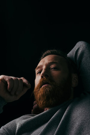 Low angle view of stylish bearded man in jumper looking at camera isolated on black with shadowの写真素材