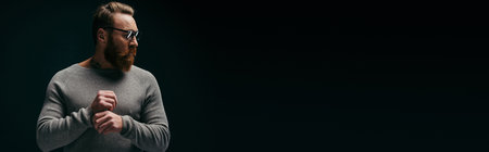 Trendy bearded man in jumper posing and looking away isolated on black, bannerの写真素材