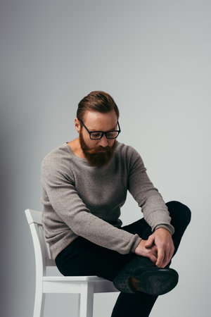 Trendy bearded man in eyeglasses touching leg while sitting on chair on grey backgroundの写真素材