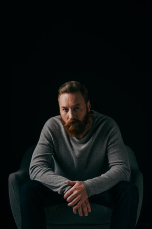 Fashionable man in jumper looking at camera near armchair isolated on blackの写真素材