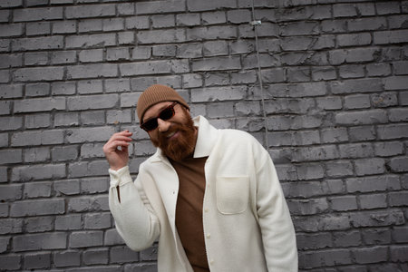 happy and bearded man in beanie hat and sunglasses smiling near brick wall