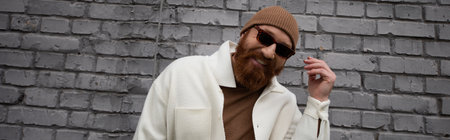 happy and bearded man in beanie hat and sunglasses smiling near brick wall, banner