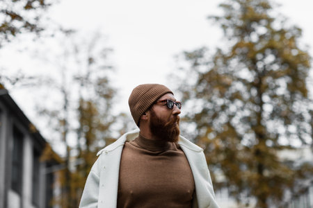 stylish bearded man in beanie hat and trendy sunglasses looking away against trees and cloudy skyの写真素材