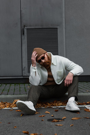 full length of stylish hipster in beanie hat and sunglasses sitting on road border on street