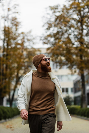 cheerful bearded man in beanie hat and trendy sunglasses looking away against trees and cloudy skyの写真素材