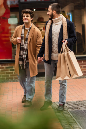 bearded gay man with shopping bags holdings hands with happy boyfriend while walking near blurred shop on streetの写真素材