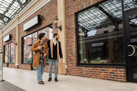 full length of trendy gay men with shopping bags and paper cup walking near showcases