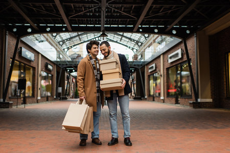 full length of trendy gay couple with shopping bags and shoeboxes near building with stores on urban streetの写真素材