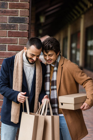 smiling homosexual couple in stylish outfit looking into shopping bags near brick column on city streetの写真素材