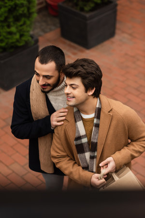 high angle view of joyful and trendy gay couple with shopping bags looking away on street