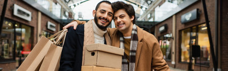 cheerful man with shopping bags embracing gay partner with shoeboxes near blurred shops on background, bannerの写真素材