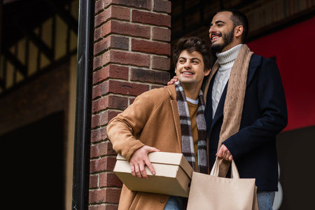 cheerful and stylish gay couple with shopping bags and shoebox looking away near brick columnの写真素材