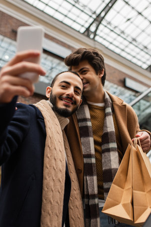 cheerful bearded man taking selfie with boyfriend holding shopping bags outdoorsの写真素材