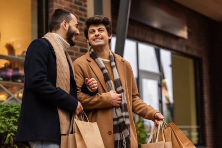trendy gay men in scarfs holding shopping bags and smiling on city streetの写真素材