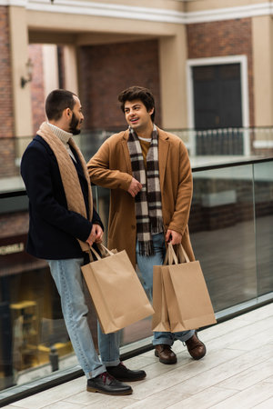 full length of fashionable gay couple with shopping bags talking near glass fence on urban streetの写真素材
