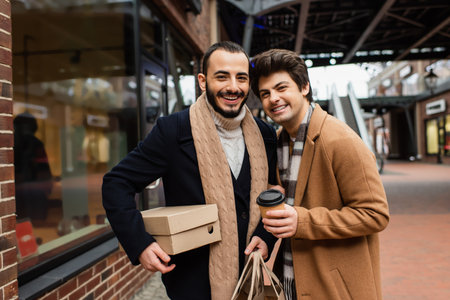 happy and fashionable gay couple with purchases and coffee to go looking at camera near showcase on streetの写真素材