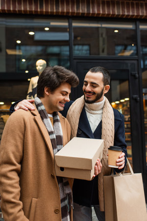 happy gay man holding shoebox near bearded boyfriend with paper cup and shopping bag at blurred showcaseの写真素材