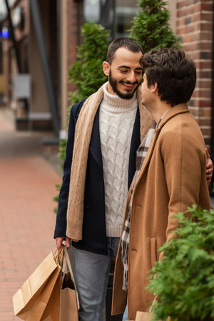 bearded gay man in trendy outfit holding shopping bags and smiling near boyfriend on streetの写真素材