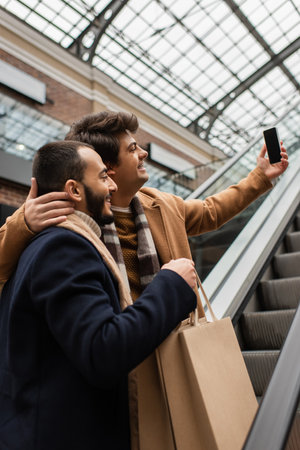young man hugging neck of bearded and trendy boyfriend with shopping bags and taking selfie on escalatorの写真素材