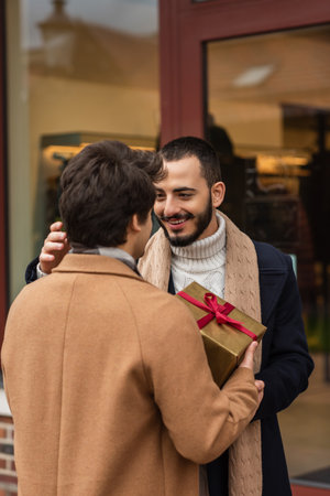 pleased bearded gay man smiling near boyfriend with Christmas gift box and blurred showcaseの写真素材