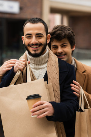 cheerful gay man embracing bearded boyfriend holding paper cup and shopping bagsの写真素材