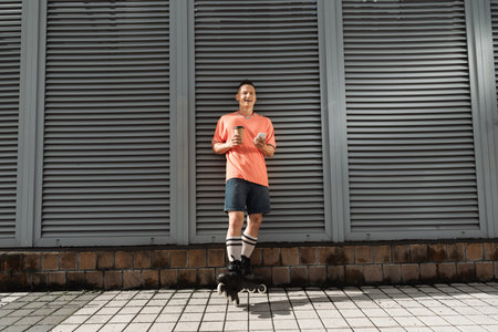 Smiling man in roller skates holding mobile phone and coffee to go near building on street at...
