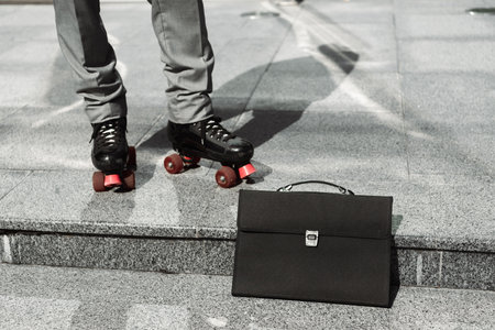 black briefcase near cropped businessman in roller skates on sidewalk of city streetの写真素材