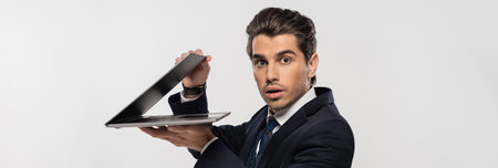 stunned businessman in suit looking at camera and holding laptop isolated on grey, banner