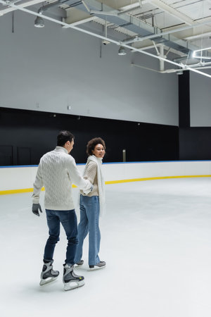 Cheerful african american woman holding hand of boyfriend while ice skating on rinkの写真素材