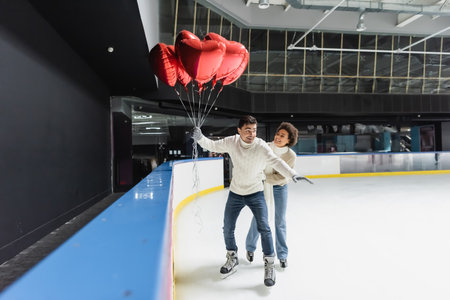 Cheerful african american woman in warm clothes ice skating near boyfriend with heart shaped balloons on rink